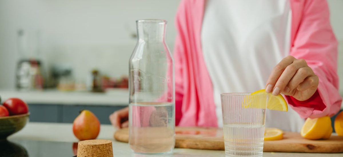 A woman in a pink outfit prepares lemon water in a bright, modern kitchen setting.